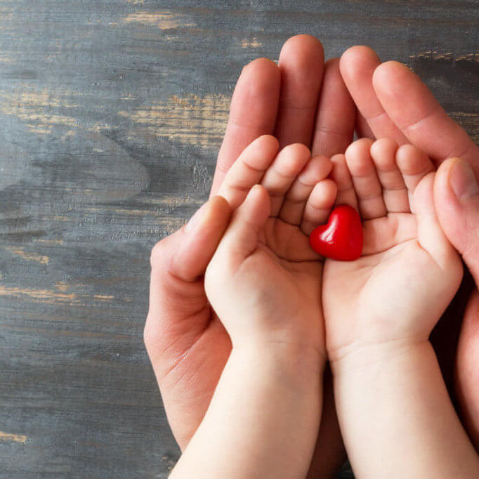Two pairs of hands, men and children on a wooden background. They hold a tiny red heart in their hands. Conception : mettre toutes les chances de son côté